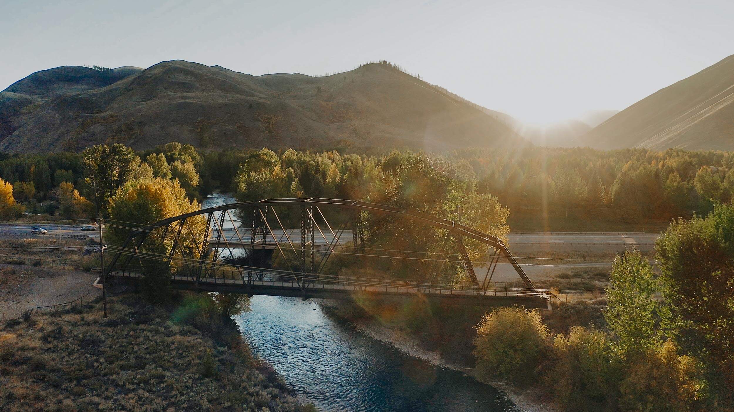 side view of a bridge crossing over a river lined with trees with mountains in the background