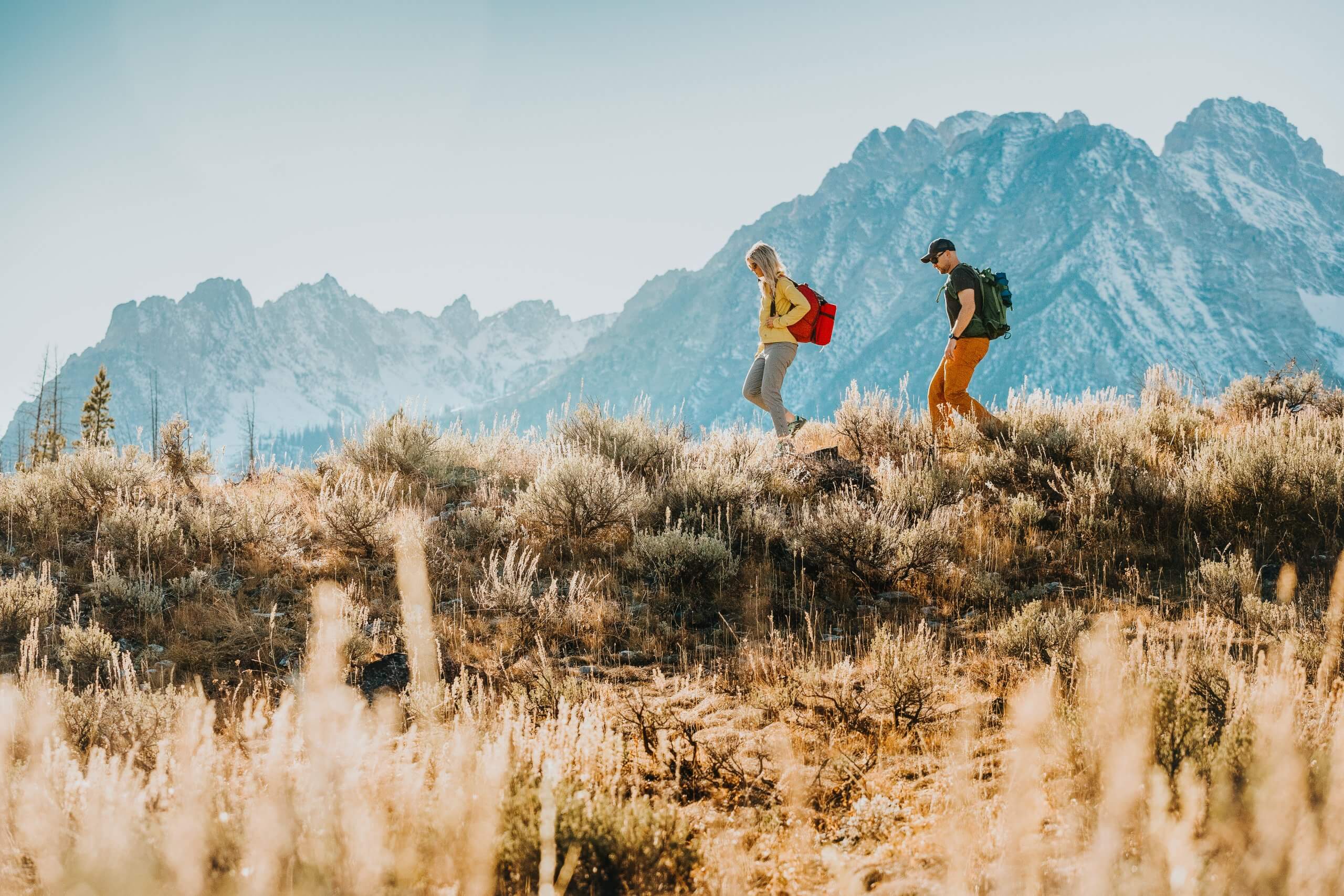 a man and woman hiking with backpacks surrounded by desert shrubs with mountains in the background