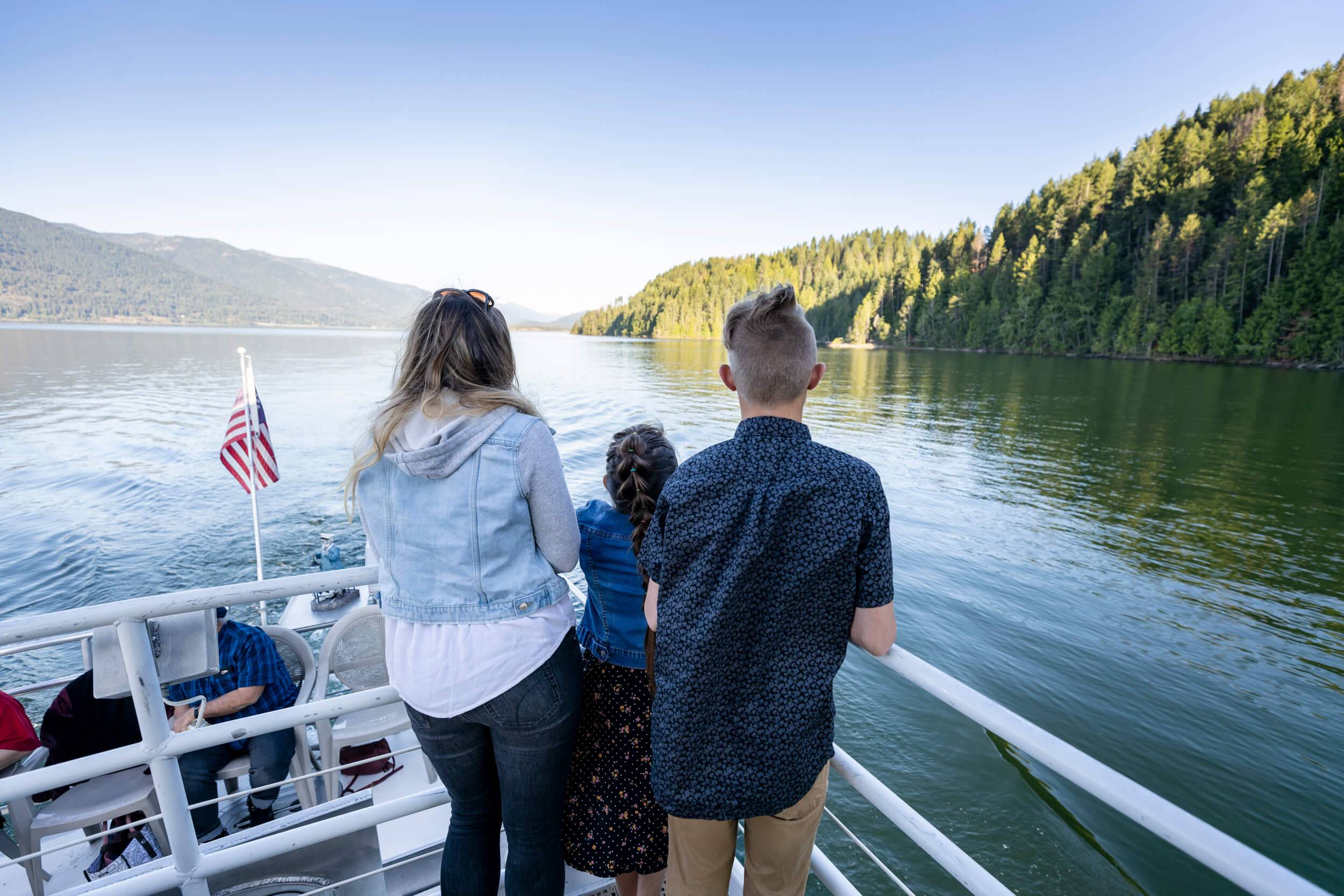 Three people stand at the edge of a boat, overlooking a lake.