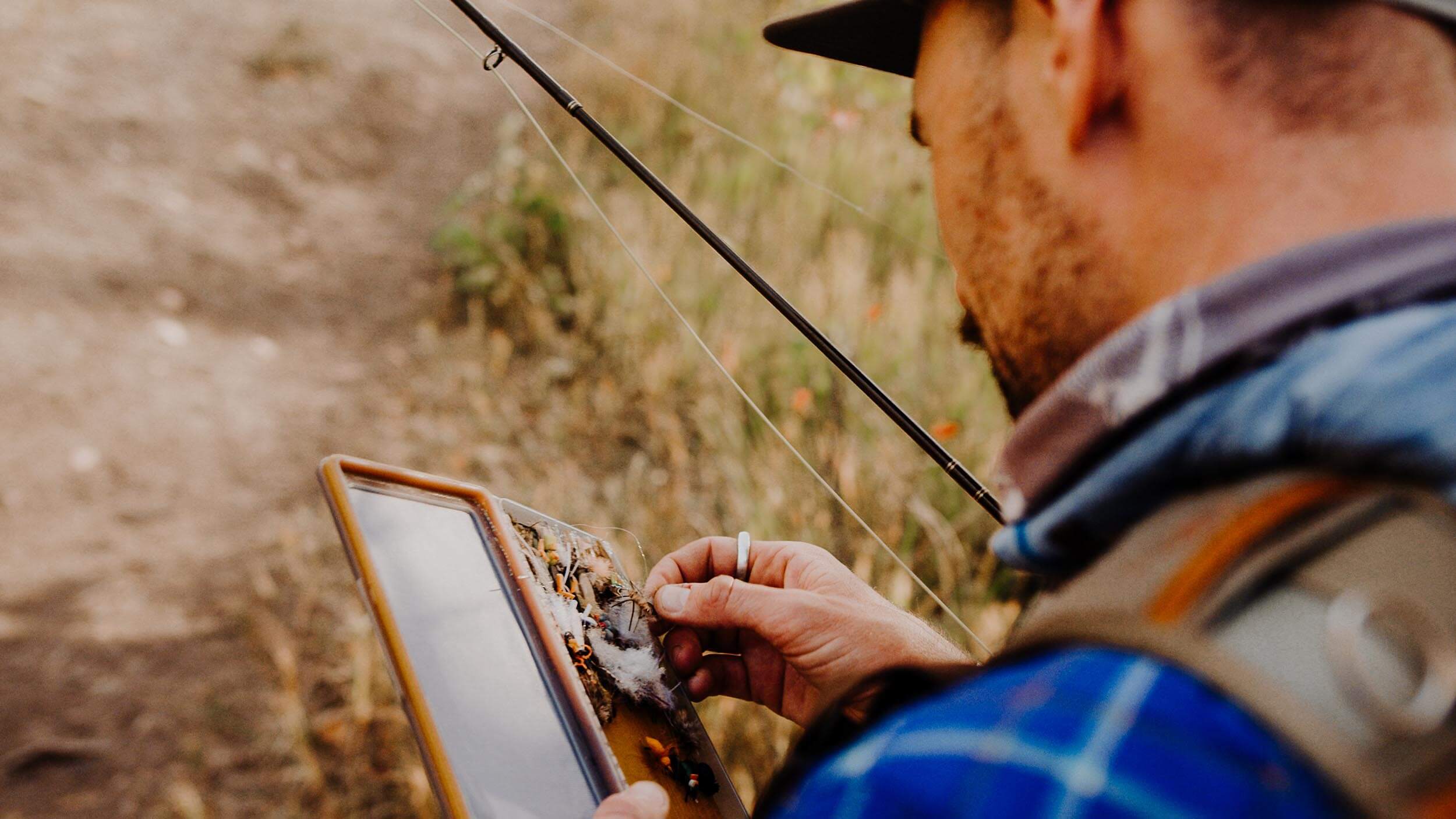 a man with a fishing rod, selecting a fishing lure from a small case