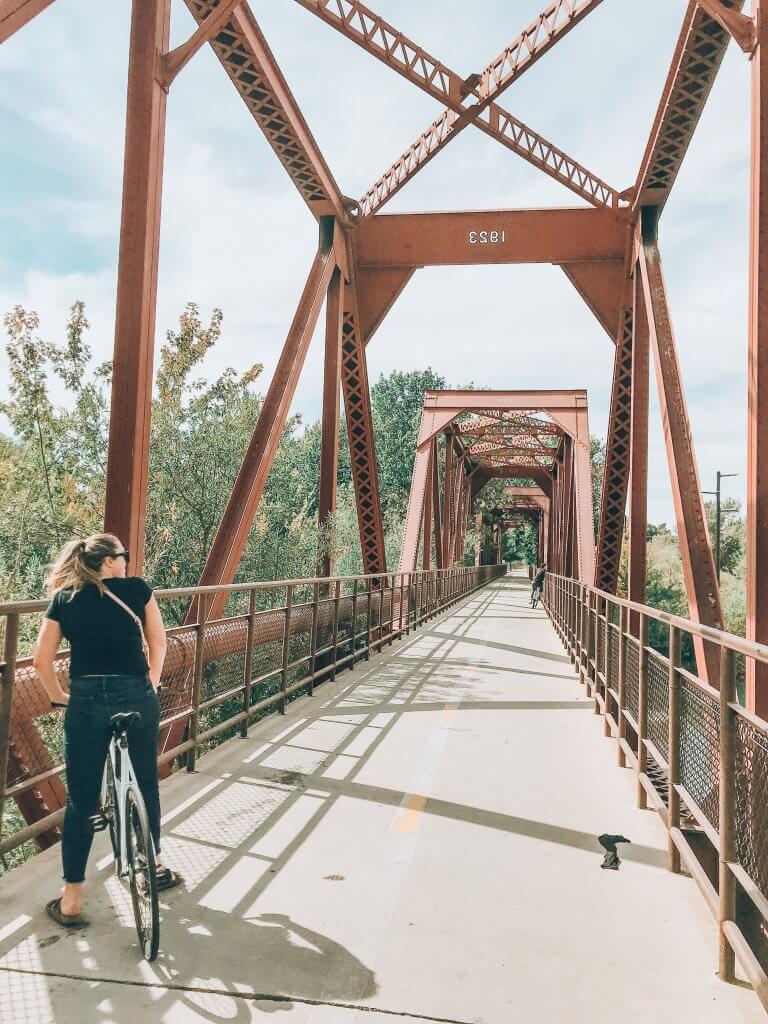 A woman stands over the top of her bicycle at the entrance to a large bridge with steel beams stretching over the top.