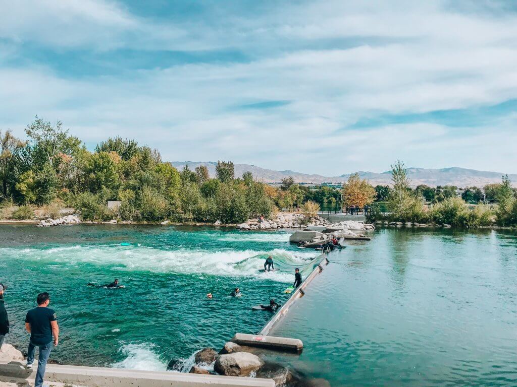 Surfers in wet suits sit in the water of the Boise River at the artificial wave waiting to surf.