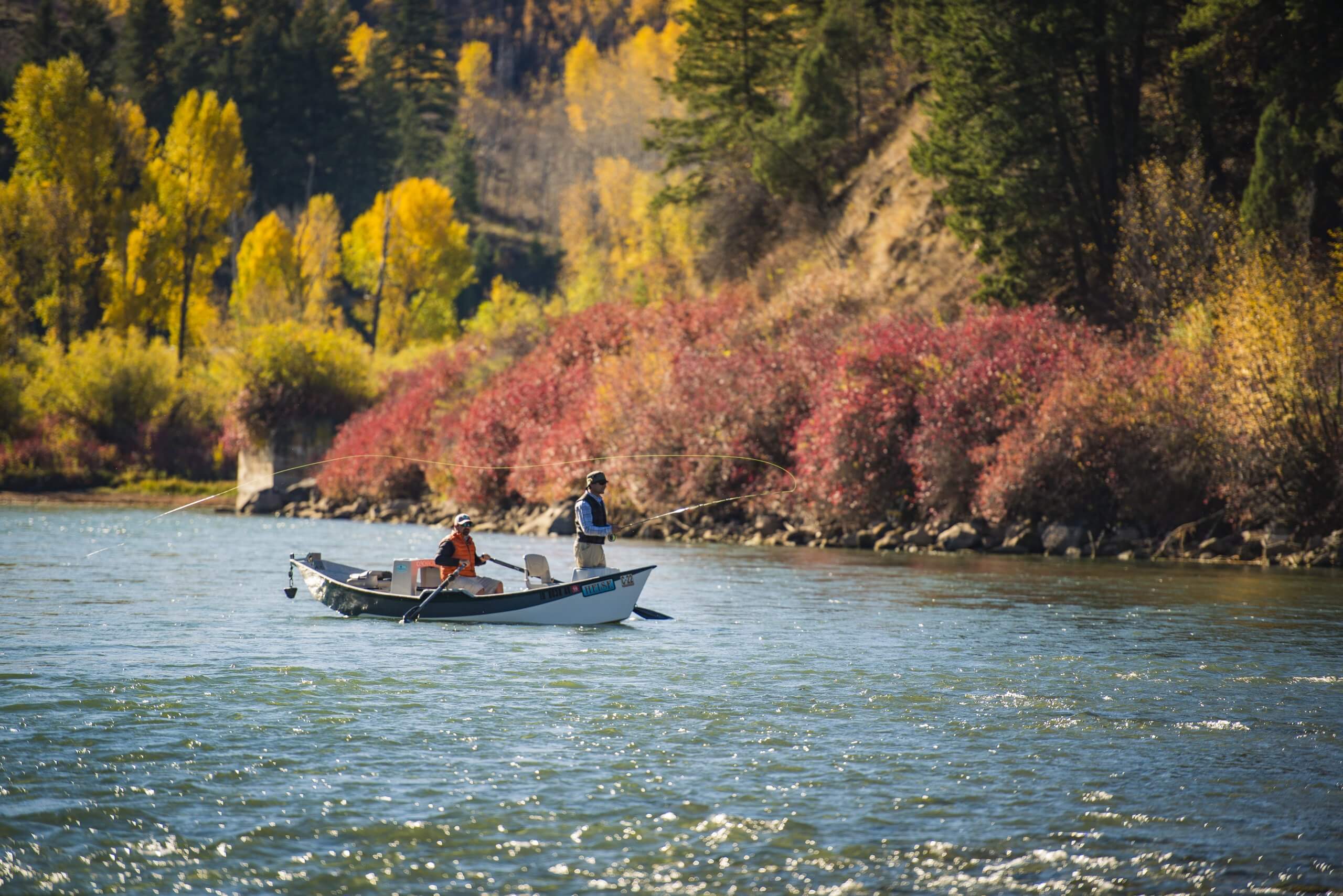 Two men cast lines from a drift boat in the South Fork of the Snake River, surrounded by fall colors.