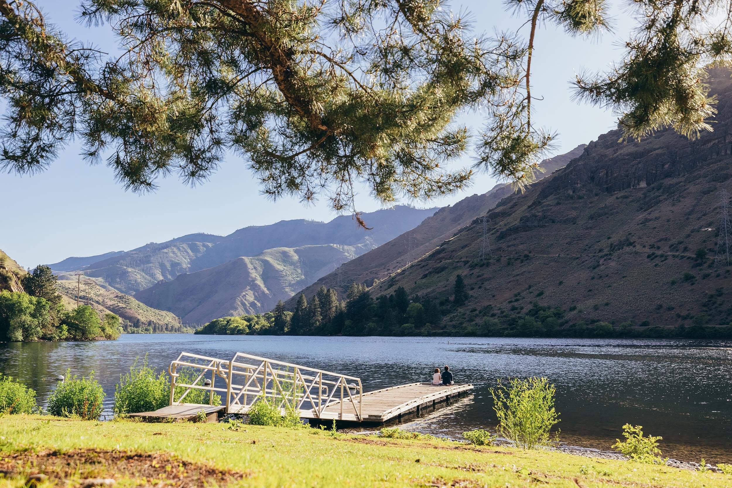 two people sitting on the edge of a dock with mountains in the background