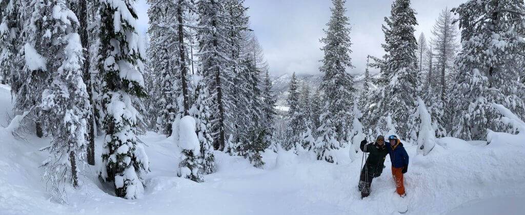 Two skiers in snow gear standing next to each other outside surrounded by deep drifts and snow-covered trees.