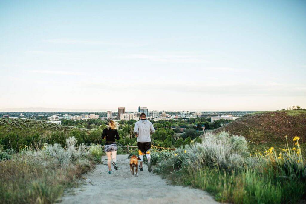 a man and woman running with their dog on a trail with Boise in the distance.