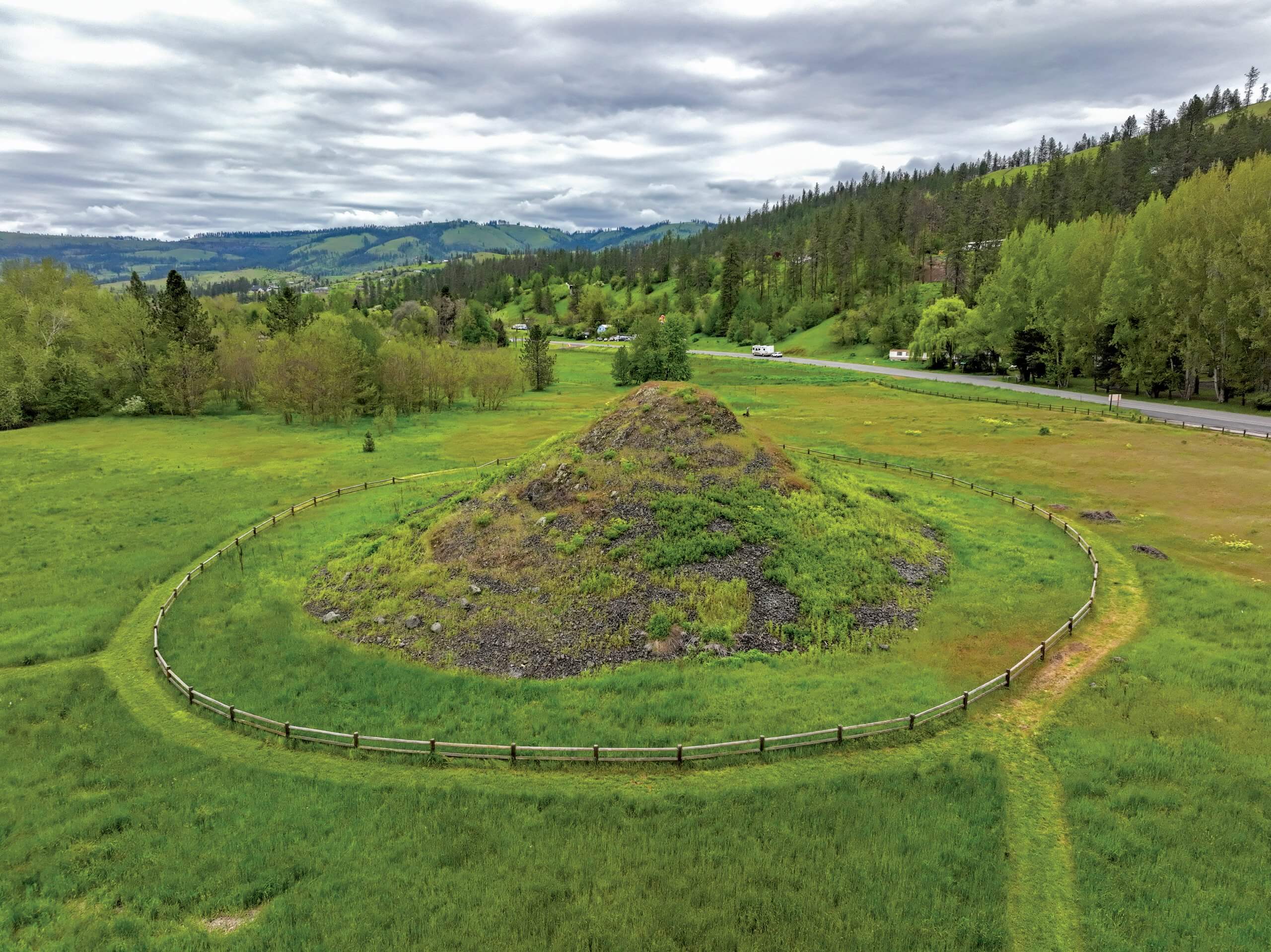 An aerial view of the Heart of the Monster site, surrounded by a sprawling field and a tree-covered hill off the side.