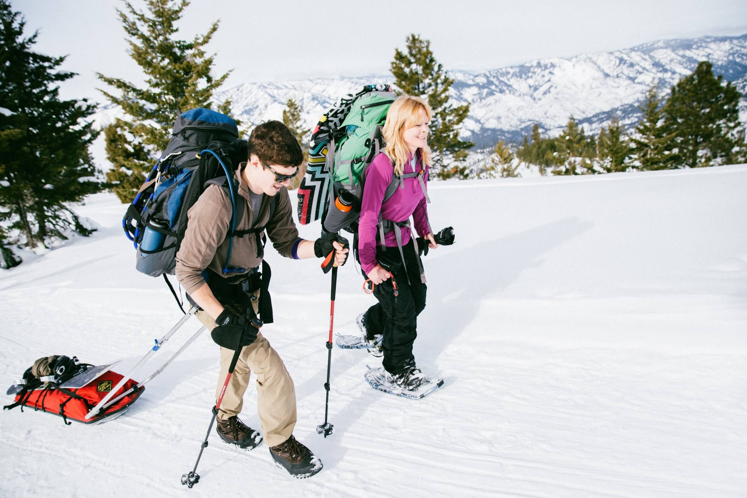 Two people with large backpacks snowshoeing up a hill, with snow-covered mountains in the distance.